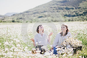 Two young pretty women having picnic with tea in chamomile field