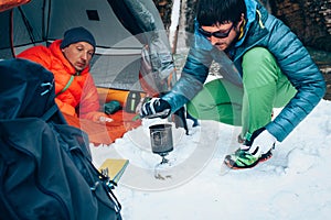 Two young professional male tourists are preparing food and hot drinks in the mountains near the river in winter.
