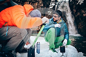 Two young professional male tourists are preparing food and hot drinks in the mountains near the river in winter.