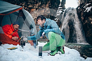 Two young professional male tourists are preparing food and hot drinks in the mountains near the river in winter.