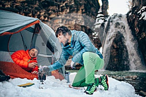 Two young professional male tourists are preparing food and hot drinks in the mountains near the river in winter.