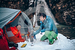 Two young professional male tourists are preparing food and hot drinks in the mountains near the river in winter.