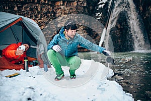 Two young professional male tourists are preparing food and hot drinks in the mountains near the river in winter.