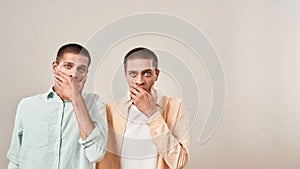 Two young scared twin brothers covering mouth with hand and looking at camera with shocked face expression while