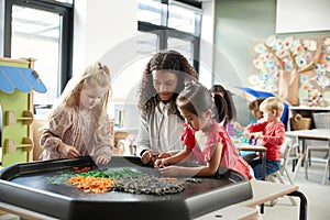 Two young schoolgirls standing at a table playing a game with their female teacher in an infant school classroom, selective focus