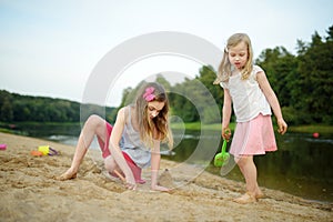 Two young sisters having fun on a sandy lake beach on warm and sunny summer day. Kids playing by the river