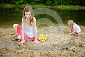 Two young sisters having fun on a sandy lake beach on warm and sunny summer day. Kids playing by the river