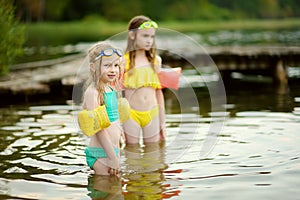 Two young sisters having fun on a sandy lake beach on warm and sunny summer day. Kids playing by the river