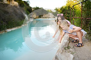 Two young sisters playing by natural swimming pool in Bagno Vignoni, with thermal spring water and waterfall. Tuscany, Italy