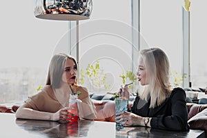 Two young smart beautiful girls in dresses sit at a restaurant table opposite a large window. Drink red and blue