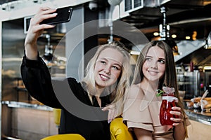 Two young smart beautiful girls in dresses are sitting at the bar. Drink red and blue cocktails from glasses with