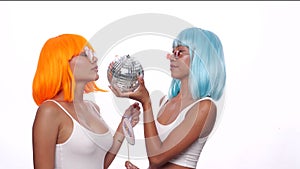 Two young smiling Asian women in bright color wigs posing with photo booth props and small disco ball over white