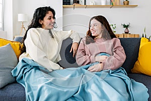 Two young smiling latin american women covered with a blanket talking on sofa