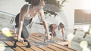 Two young smiling women, girls in sportswear doing exercises while listening to music. Workout, couching on city street.