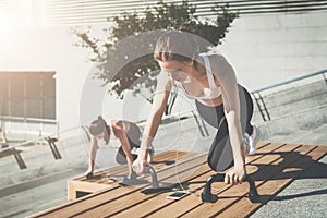 Two young smiling women, girls in sportswear doing exercises while listening to music. Workout, couching on city street.