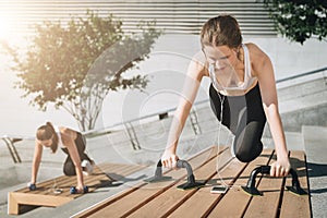 Two young smiling women, girls in sportswear doing exercises while listening to music. Workout, couching on city street.