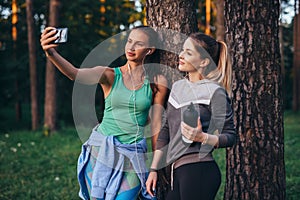 Two young sportive girlfriends wearing sportswear leaning against tree taking selfie with smartphone in forest