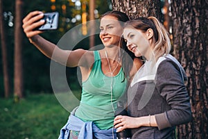 Two young sportive girlfriends wearing sportswear leaning against tree taking selfie with smartphone in forest