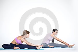 Two young sports women doing stretching exercises while sitting on a yoga mat on a white