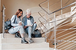 Two young student friends sitting and reading book together on the stairs in college