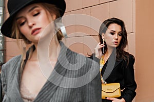 Two young stylish beautiful women fashion models are posing in street, wearing pantsuit, hat, having purse on waist.