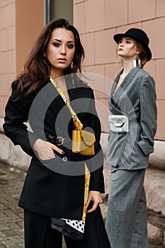 Two young stylish beautiful women fashion models are posing in street, wearing pantsuit, hat, having purse on waist.