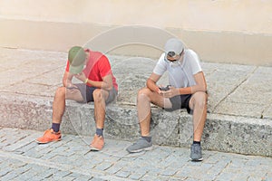 Two young teenagers sitting on the stone pavement playing and chatting on social networks with their mobile phone with their