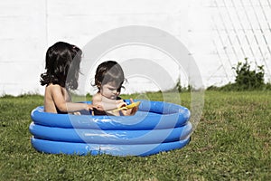 Two young toddler female sisters playing on a blue small paddling pool in the grass