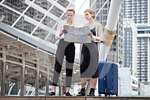Two young tourists women looking at map and searching destination in urban city outdoors . friends traveling with luggage together
