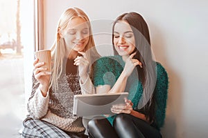 Two young woman chatting in a coffee shop. Two friends enjoying coffee together. One girl uses a tablet.