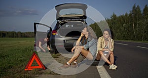Two young woman sit while man repair broken car