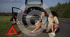 Two young woman sit and use phone to find help while man repair broken car