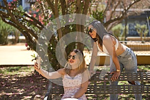 Two young women, beautiful, brunette and blonde, South Americans with sunglasses, sitting on a bench taking pictures with their