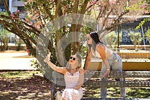 Two young women, beautiful, brunette and blonde, South Americans with sunglasses, sitting on a bench taking pictures with their