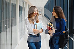 Two young women with book chatting while standing in college corridor. University students in corridor after the lecture