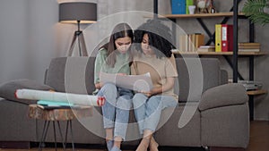 Two young women entering living room sitting down on couch analyzing business graph. Portrait of concentrated Asian and