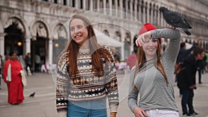 Two young women feed the pigeons from their hands in the city square of Venice.