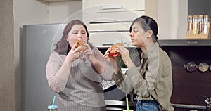 Two young women friends eating juicy burgers and chatting in the kitchen.