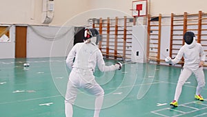 Two young women in full protection of white suits having an active fencing training in the school gym