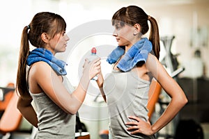 Two young women in grey sportswear with towels standing with bottle of water and looking at each other in gym