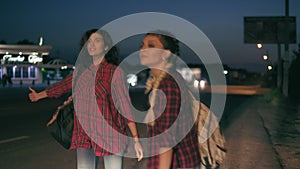 Two young women hitchhikers stand on the sidelines holding their backpacks and ask to stop the passing cars in the night