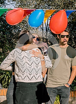 Two young women hugging at a surprise birthday party and a boy next to them smiling. Vertical view