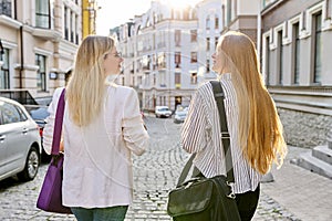 Two young women with laptop bags walking along street of sunset city, back view