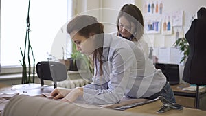 Two young women lay out cloth on table in sewing workshop, female professionals are in working process with fabric, near