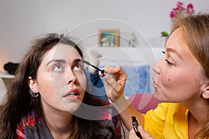 Two young women lie on the bed and make up decorative cosmetics. One woman paints another woman`s eyelashes with mascara. Close u