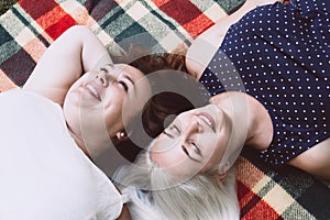 Two young women lie on bright plaid blanket in nature