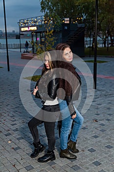 Two young women with long hair and black leather jackets are friends in park against the background of neon sign of coffee shop