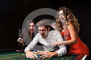 Two young women and man behind roulette table on black background