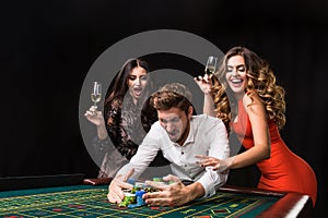 Two young women and man behind roulette table on black background