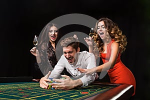 Two young women and man behind roulette table on black background